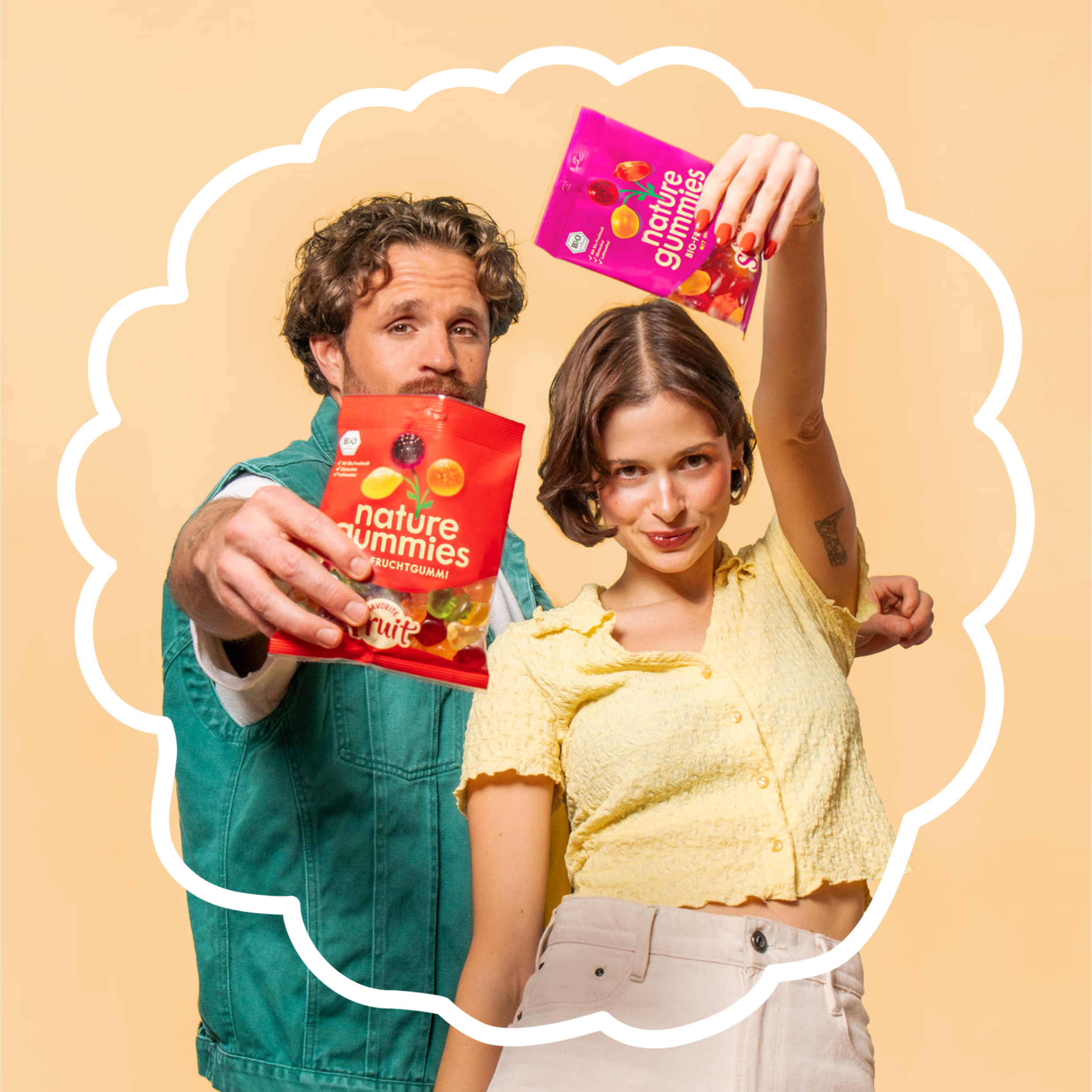 Ein junger Mann mit lockigen Haaren und eine junge Frau mit kurzen, dunklen Haaren präsentieren fröhlich zwei bunte Packungen nature gummies. Die Frau hebt eine violette Simply Super Packung in die Luft, während der Mann eine rote Packung Favorite Fruits nach vorne hält. Beide lächeln spielerisch vor einem beigefarbenen Hintergrund, umrahmt von einer weißen, Brombeer-förmigen Kontur.