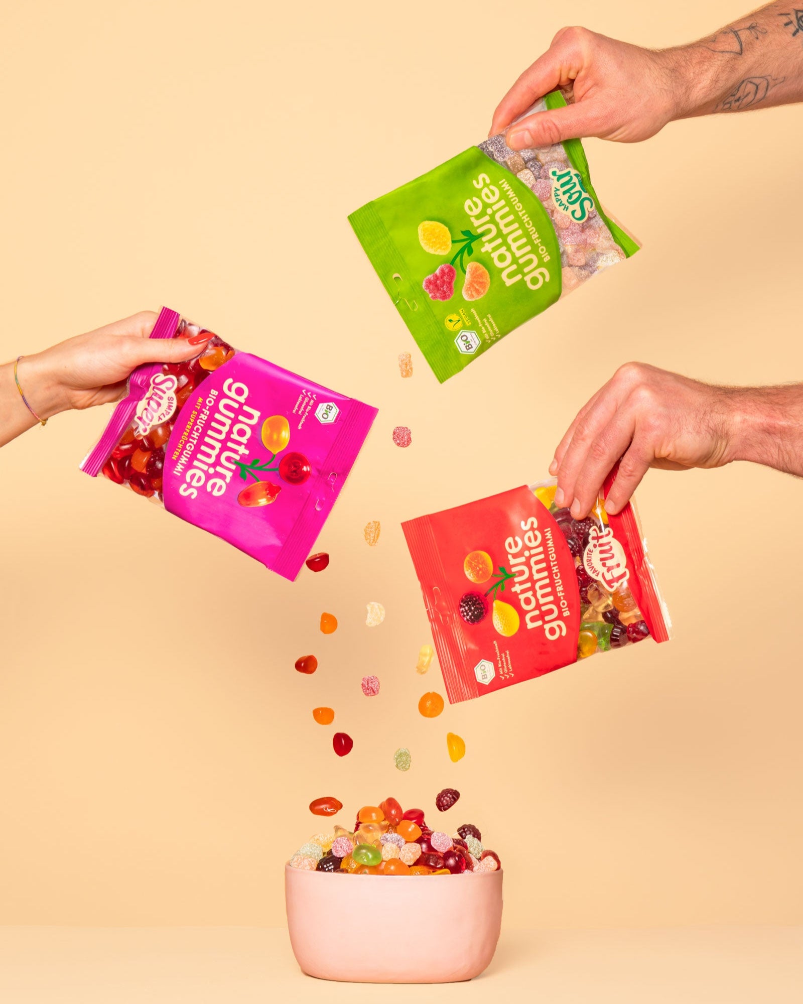 Drei Hände halten bunte nature gummies Packungen und schütten den Inhalt in eine rosa Schüssel. Die Gummibärchen fallen in verschiedenen Farben und Formen vor einem beigefarbenen Hintergrund.