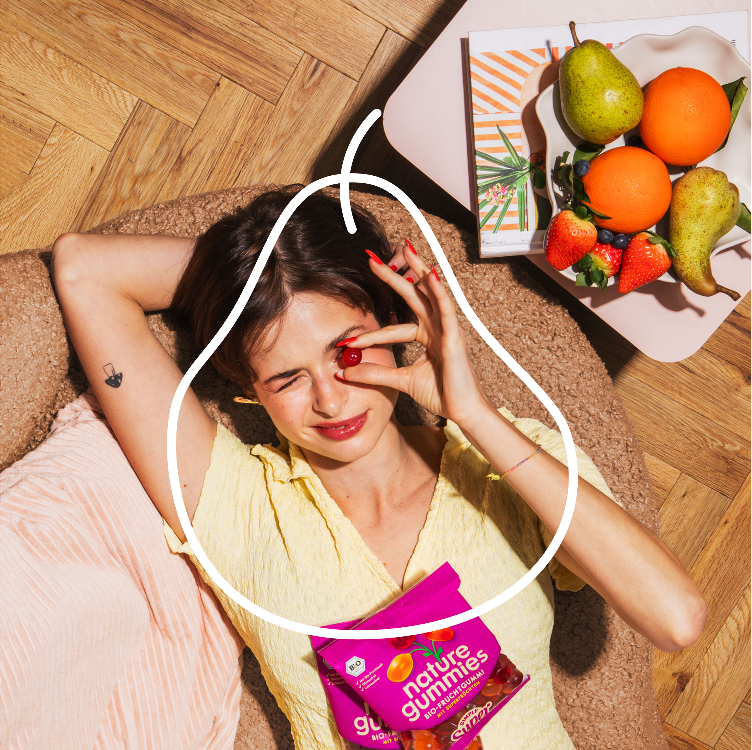 Eine Frau entspannt auf einer Couch und hält sich ein Fruchtgummi der Sorte Simply Super direkt vor ihr Auge, sodass es so aussieht, als wäre es ihr Auge. Neben ihr steht eine frisch und fruchtig anmutenden Obstschale auf einem Beistelltisch.