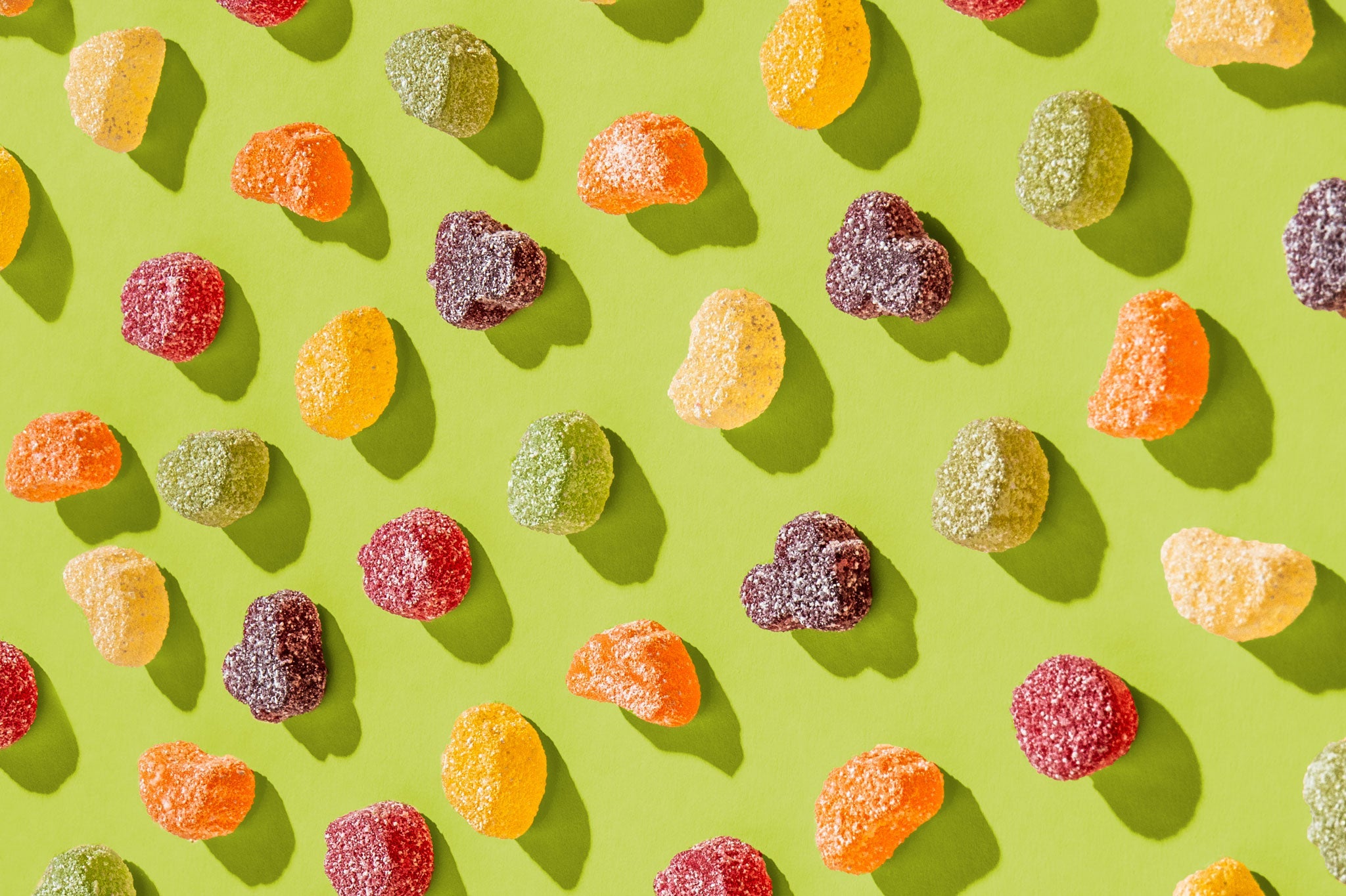Bunte, gezuckerte Fruchtgummis in verschiedenen Farben und Formen sind gleichmäßig auf einem grünen Hintergrund angeordnet. Die Gummis werfen durch das Licht weiche Schatten, wodurch ihre Struktur und der Zuckerüberzug betont werden.