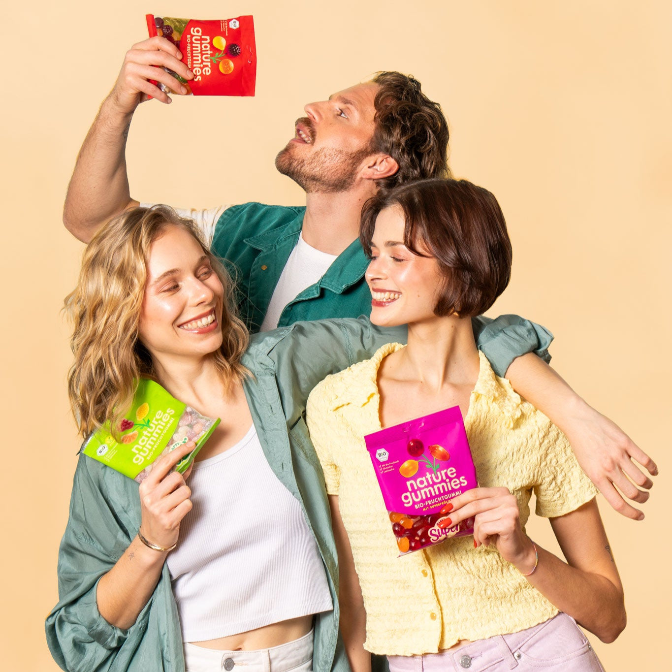 Drei junge Menschen genießen fruchtige nature gummies vor einem beigefarbenen Hintergrund. Der Mann in der Mitte trägt eine grüne Weste und hält eine rote Packung hoch, während er die Gummibärchen isst. Die beiden Frauen an seiner Seite lachen und schauen sich gegenseitig an – eine hält eine grüne Packung, die andere eine pinke. Die Szene wirkt lebendig und ausgelassen.