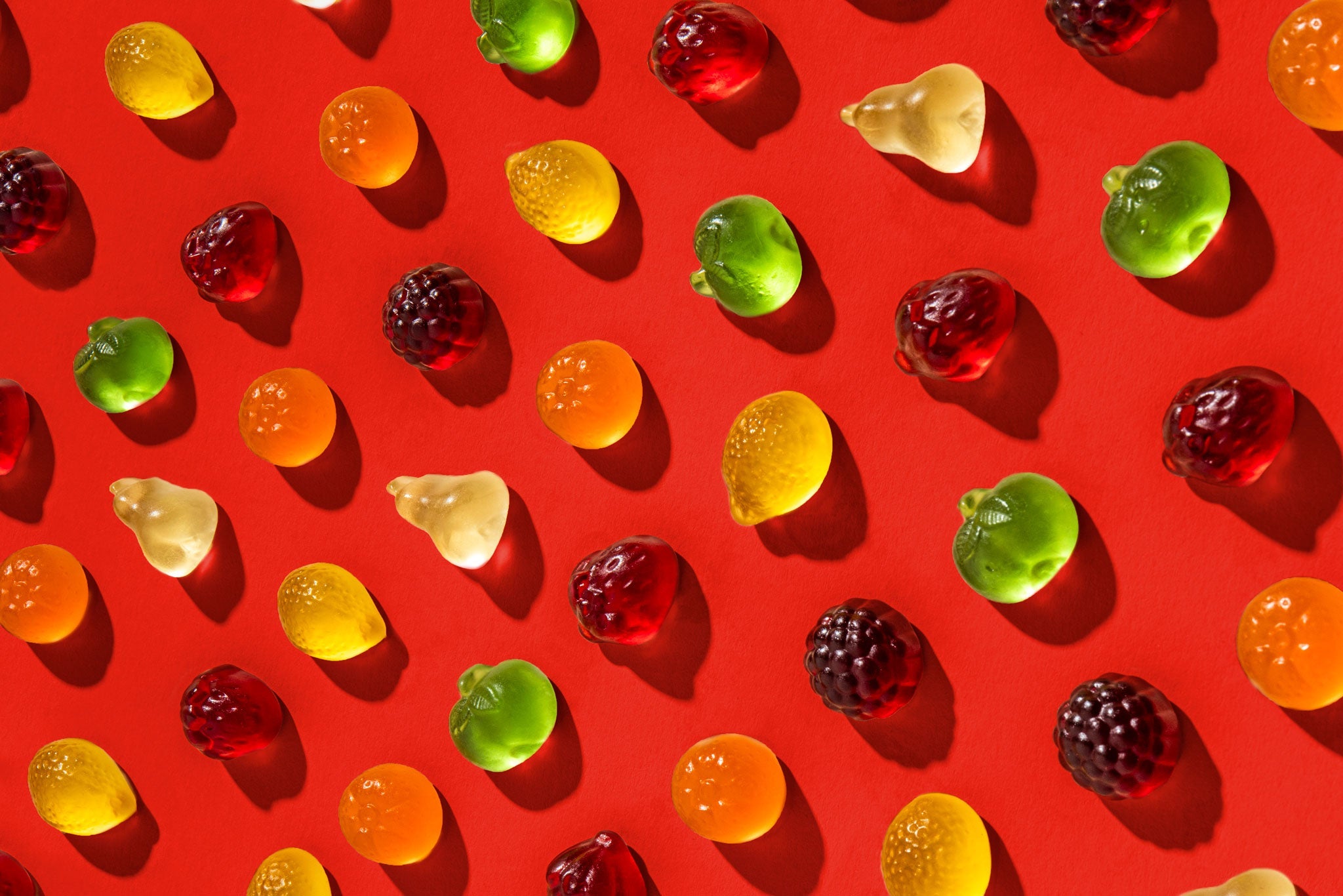 Bunte Fruchtgummis in Form verschiedener Früchte, darunter Brombeeren, Äpfel, Birnen und Zitronen, Orangen und Erdbeeren, sind auf einem roten Hintergrund gleichmäßig angeordnet. Die Fruchtgummis werfen kleine, weiche Schatten und leuchten durch das Licht, was ihre Transparenz betont.