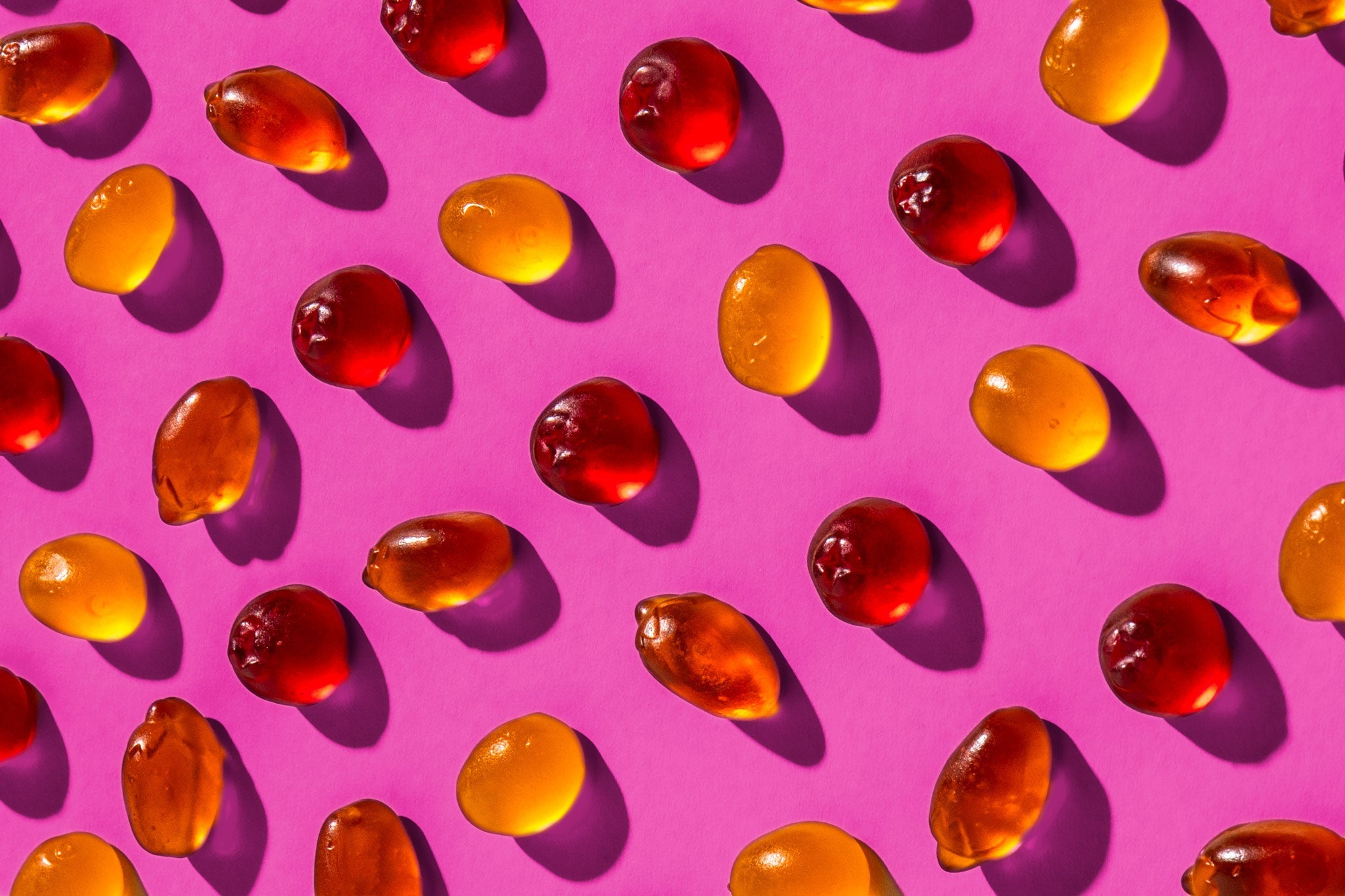 Halbtransparente Fruchtgummis in warmen Rot- und Gelbtönen sind gleichmäßig auf einem leuchtend pinken Hintergrund angeordnet. Das Licht erzeugt sanfte Schatten, die die glänzende Oberfläche und die weiche Struktur der Gummis betonen.